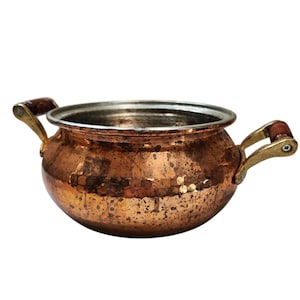 May include: A hammered copper pot with brass handles and wooden grips. The pot has a wide, open mouth and a silver-colored rim. The copper has a rich, warm color with a textured surface. The handles are attached to the pot with brass hardware.