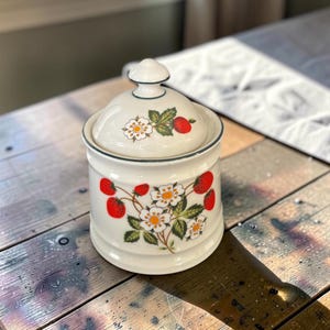 May include: A cream-coloured ceramic canister with a matching lid, featuring a botanical pattern of red strawberries, green leaves, and white flowers with yellow centres. The canister has a dark green rim accent on both the lid and the body.