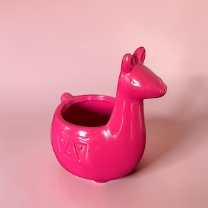 Puede incluir: Una maceta rosa brillante con forma de llama. La maceta de cerámica presenta un cuerpo redondo, una cara detallada, orejas y una pequeña cola. La maceta tiene un patrón decorativo alrededor de la base y está sobre un fondo rosa.