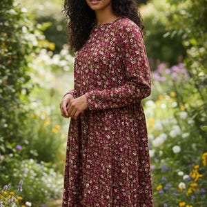 Può includere: Abito bordeaux a maniche lunghe con fantasia floreale. L'abito presenta uno scollo tondo e una gonna fluida. La stampa floreale include accenti bianchi, rosa e verdi. L'abito è indossato all'aperto in un giardino.