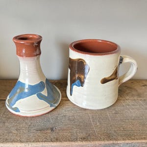 May include: A handmade ceramic mug and vase set. The mug is off-white with brown and blue abstract designs. The vase is white with blue and brown abstract designs. Both pieces have a brown interior and are on a wooden surface.