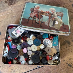 May include: A rectangular metal storage tin filled with a large assortment of sewing buttons in various colors, sizes, and materials. The lid features a printed illustration of two figures in period clothing playing a game at a wooden table.