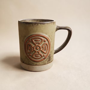 May include: A ceramic mug with a cylindrical shape, featuring a raised, circular design in shades of orange and brown. The mug has a light green body, a dark brown handle, and a cream-colored base. The mug is approximately 4 inches tall.