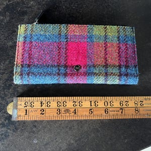 May include: A rectangular wallet with a plaid pattern in shades of blue, pink, green, and red. The wallet has a small metal emblem with the letter "M" on the front. The wallet is approximately 4 inches wide.