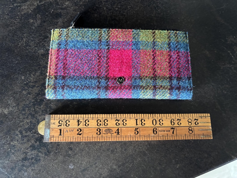 May include: A rectangular wallet with a plaid pattern in shades of blue, pink, green, and red. The wallet has a small metal emblem with the letter "M" on the front. The wallet is approximately 4 inches wide.