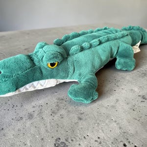 May include: A green plush alligator toy with a detailed design. The alligator has a textured body, yellow eyes, and a white mouth with grey teeth. The toy is lying on a grey surface.