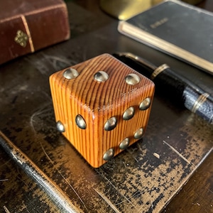 Könnte beinhalten: Ein Würfel aus Holz mit Messingnieten, die die Zahlen markieren. Der Würfel besteht aus hellfarbigem Holz mit sichtbarer Maserung und liegt auf einer dunklen, abgenutzten Oberfläche. Ein Stift und ein Notizbuch sind im Hintergrund.