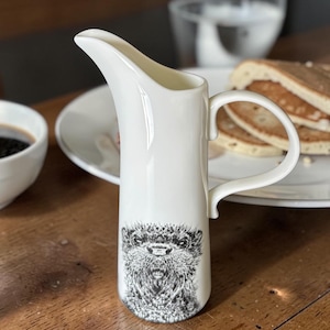 May include: A white ceramic creamer with a black and white illustration of an otter. The creamer has a curved handle and a spout for easy pouring. The creamer is on a wooden table.