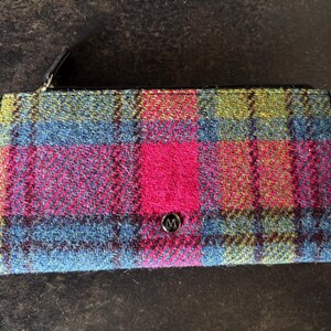 May include: A rectangular wallet with a colorful plaid pattern in shades of blue, pink, green, and red. The wallet has a black zipper and a small, round, silver button with the letter "M" on it.