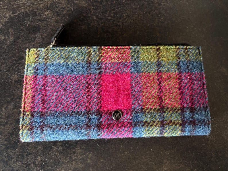 May include: A rectangular wallet with a colorful plaid pattern in shades of blue, pink, green, and red. The wallet has a black zipper and a small, round, silver button with the letter "M" on it.