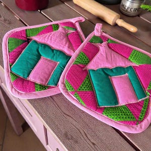 May include: A pair of quilted pot holders with a pink, green, and magenta geometric pattern. Each has a house-shaped design in pink and green, a pink border, and a hanging loop. The pot holders are for kitchen use.