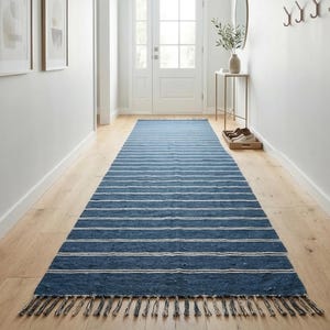 May include: A long, rectangular blue and white striped rug with a fringed edge. The rug is placed in a hallway with light wood flooring and white walls. The rug's design features horizontal stripes.
