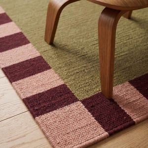 Op de afbeelding: Een geweven vloerkleed met een groot olijfgroen middenpaneel, omrand door een herhalend geblokt patroon van afwisselende oudroze en donkerbordeauxrode vierkanten. Een gebogen houten stoelpoot rust op het kleed, dat het BLR-logo bevat.