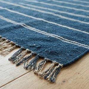 May include: A blue and white striped rug with a fringed edge. The rug has a woven texture and is made of a durable material. The rug is placed on a light-colored wooden floor.