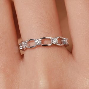 Op de afbeelding: Een zilverkleurige ring met een schakelontwerp met kleine, heldere, rond geslepen stenen in afwisselende schakels. De ring heeft een gepolijste metalen afwerking en een doorlopend patroon van met elkaar verbonden ovale metalen segmenten.