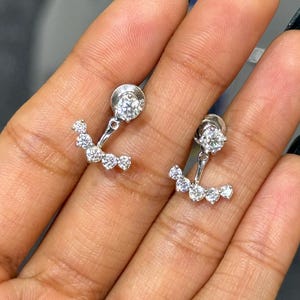 May include: A pair of silver-toned ear jackets featuring a single round-cut clear stone stud at the top, connected to a curved, crescent-shaped bar set with five smaller round-cut clear stones that hang below the earlobe when worn.