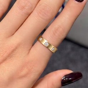May include: A gold band ring featuring a series of small, square-cut clear stones set flush into the metal surface. The ring is worn on the ring finger, which is positioned alongside other fingers displaying dark, glossy, deep burgundy nail polish.