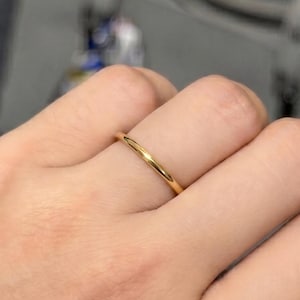 Può includere: Un semplice anello a fascia in oro lucido con una superficie liscia e arrotondata. L'anello presenta un design classico e minimalista con una finitura metallica brillante, indossato su un dito. La fascia ha larghezza e spessore uniformi.
