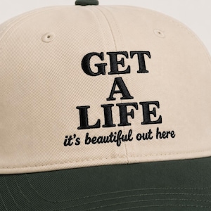May include: A beige baseball cap with a dark green brim and a dark green button on top. The front features black embroidered text that reads GET A LIFE in a bold serif font, followed by the text it's beautiful out here in a smaller script font.