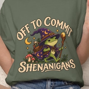 May include: An olive green t-shirt featuring a graphic of a green frog wearing a purple wizard hat and cape, carrying a staff and a backpack. The text reads "OFF TO COMMIT SHENANIGANS" in a curved font, surrounded by stars, a crescent moon, and mushrooms.