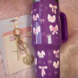 Puede incluir: Un vaso morado con asa y tapa transparente, decorado con lazos y corazones rosas. Un llavero dorado con una cruz y un colgante en forma de corazón está junto al vaso. El llavero está sujeto a una tarjeta que dice "ENCHANTÉ."