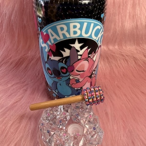 May include: A clear glass tumbler with a black interior and a Starbucks logo featuring Stitch and Angel. A wooden honey dipper with a jeweled end rests on a clear, faceted glass ashtray. The items are on a pink, furry surface.