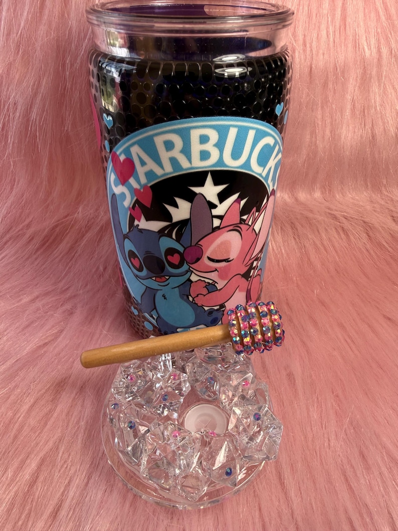 May include: A clear glass tumbler with a black interior and a Starbucks logo featuring Stitch and Angel. A wooden honey dipper with a jeweled end rests on a clear, faceted glass ashtray. The items are on a pink, furry surface.