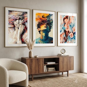 Puede incluir: Tres impresiones artísticas abstractas enmarcadas. La obra de arte presenta retratos con colores vibrantes y pinceladas expresivas. Los marcos son dorados y el arte se exhibe sobre un mueble de madera y una silla beige.