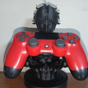 Puede incluir: Un busto negro de estilo anime sosteniendo un mando rojo de PlayStation 4. El mando es rojo con detalles y botones negros. El busto tiene cabello negro y un rostro detallado, descansando sobre una base negra. El mando está centrado.