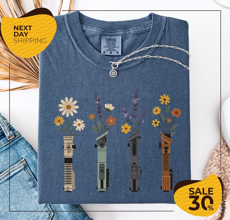 a blue t-shirt with a graphic design of four different colored vases holding flowers, arranged in a row. The vases are depicted in various colors, including blue, green, orange, and brown. The design also includes a small charm hanging from the vase. The t-shirt is displayed on a white background, and there are some jeans and a sweater visible in the background.