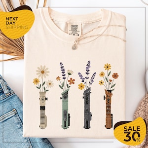 a white t-shirt with a graphic design of four colorful vases holding flowers, arranged in a row. The vases are of different colors and shapes, creating a visually appealing pattern. The t-shirt is displayed on a white background, and there are some jeans and a sweater visible in the background. The image also includes text that reads "Next Day Shipping" and "SALE 30%", indicating that the t-shirt is available for immediate shipping and is on sale at a discounted price.