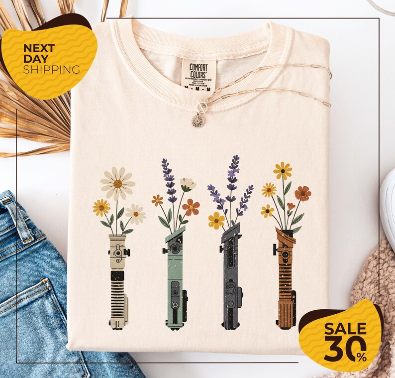 a white t-shirt with a graphic design of four colorful vases holding flowers, arranged in a row. The vases are of different colors and shapes, creating a visually appealing pattern. The t-shirt is displayed on a white background, and there are some jeans and a sweater visible in the background. The image also includes text that reads "Next Day Shipping" and "SALE 30%", indicating that the t-shirt is available for immediate shipping and is on sale at a discounted price.