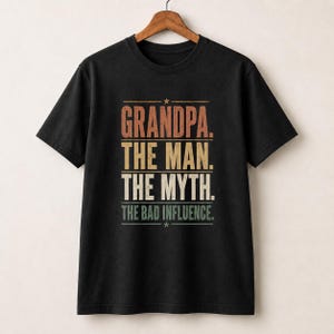 May include: A black short-sleeve t-shirt featuring a distressed, multi-colored graphic print that reads: "GRANDPA. THE MAN. THE MYTH. THE BAD INFLUENCE." The text is arranged in four horizontal lines with a small star above and below the text block.