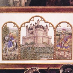 May include: A framed cross-stitch embroidery featuring a central castle on a hill flanked by two arched panels. The left panel shows a rider on a white horse, and the right panel depicts a knight on a horse. The artwork is enclosed in a brown frame.