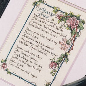May include: A framed cross-stitch embroidery featuring the lyrics to Amazing Grace in black cursive lettering. The text is bordered by a thin blue line and decorated with pink roses and green leaves in the top right and bottom left corners.