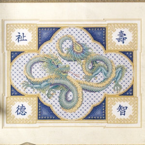 May include: A framed cross-stitch embroidery featuring a central green dragon on a blue and white patterned background. Four gold-bordered squares in the corners contain blue Chinese characters. The frame is ornate gold with a gold tassel cord.