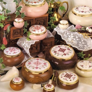 May include: A collection of round wooden and ceramic lidded containers with gold-tone rims, featuring embroidered floral patterns on the lids. The set includes various sizes, some displayed on wooden chests, accompanied by embroidery thread and scissors.