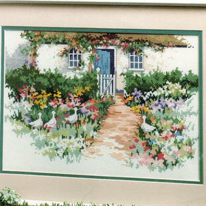 May include: A framed cross-stitch embroidery depicting a cottage with a blue door, a brick path, and a lush garden filled with colorful flowers and green bushes. Four white geese are positioned among the vibrant floral arrangements in the front yard.