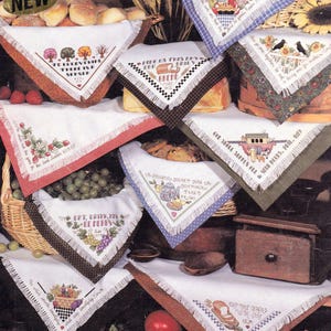 May include: A collection of white fabric bread covers with fringed edges and cross-stitched designs featuring various motifs and text, including phrases like To Everything There Is A Season, Give Us This Day Our Daily Bread, and Eat Drink And Be Merry.