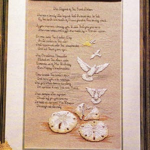 May include: A framed cross-stitch sampler on beige fabric featuring the text The Legend of the Sand Dollar in black script. The design includes embroidered white doves, a yellow star, and sand dollar illustrations in a dark wooden frame.