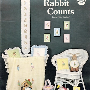 May include: A collection of Peter Rabbit Counts Beatrix Potter Creations items including a numbered wall hanging, embroidered blankets, pillows, a wicker bassinet, a wicker chair, plush rabbit and duck toys, a small basket, and framed art prints.