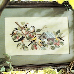 Puede incluir: Obra de arte de punto de cruz con una variedad de pájaros coloridos posados en ramas con bayas rojas y hojas verdes. También se incluye una pequeña casa para pájaros en el diseño. La obra de arte está enmarcada en un marco de madera marrón oscuro.