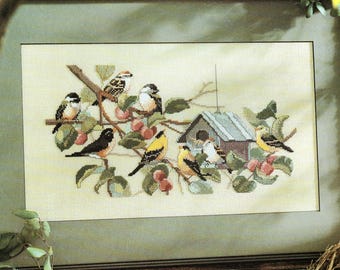 May include: Cross-stitch artwork featuring a variety of colorful birds perched on branches with red berries and green leaves. A small birdhouse is also included in the design. The artwork is framed in a dark brown wooden frame.