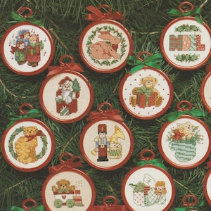 May include: A collection of round cross-stitch ornaments featuring various holiday designs including Santa Claus, a teddy bear, a nutcracker, a cat, and the word NOEL, each framed in a red plastic hoop with a small ribbon bow at the top.