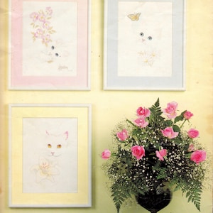 May include: Three framed prints featuring white cats with blue or yellow eyes, accented by flowers and a butterfly. A glass vase holds a large arrangement of pink roses and green ferns, with two loose pink roses resting on the surface below.