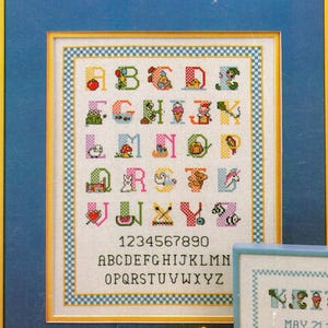 May include: A framed cross-stitch sampler featuring a colorful alphabet where each letter is decorated with small motifs like an apple, frog, owl, and unicorn. Below the alphabet are the numbers 0-9 and the alphabet in plain black capital letters.