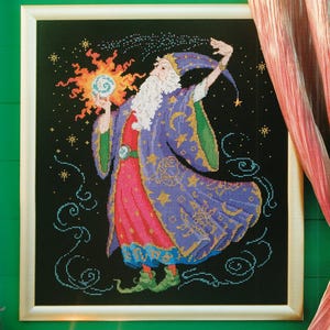 May include: Cross-stitch artwork of a wizard in a purple robe with gold accents, a red tunic, and a long white beard. The wizard holds a glowing orb and is set against a black background with stars and swirling patterns. Framed in a white wooden frame.