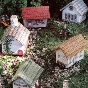 Op de afbeelding: Een verzameling miniatuurhuisjes gemaakt van naaldwerkstof, met daken in verschillende kleuren zoals rood, bruin en groen. De bouwwerken tonen gedetailleerde architecturale elementen zoals schoorstenen, ramen en uithangborden in een dorpsscène.