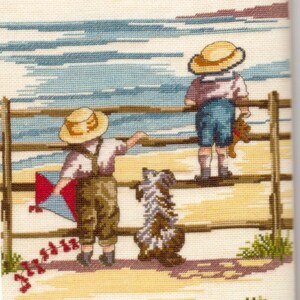 May include: A cross-stitch embroidery depicting two children in straw hats standing by a wooden fence at the beach. One child holds a red and blue kite, while the other holds a small brown toy. A shaggy dog sits between them on the sand.