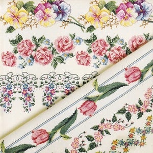 Puede incluir: Una pieza de tela blanca con varios patrones florales en punto de cruz. El diseño incluye filas horizontales de pensamientos amarillos y morados, rosas rosadas con pequeñas flores azules y una sección diagonal con tulipanes rosados.